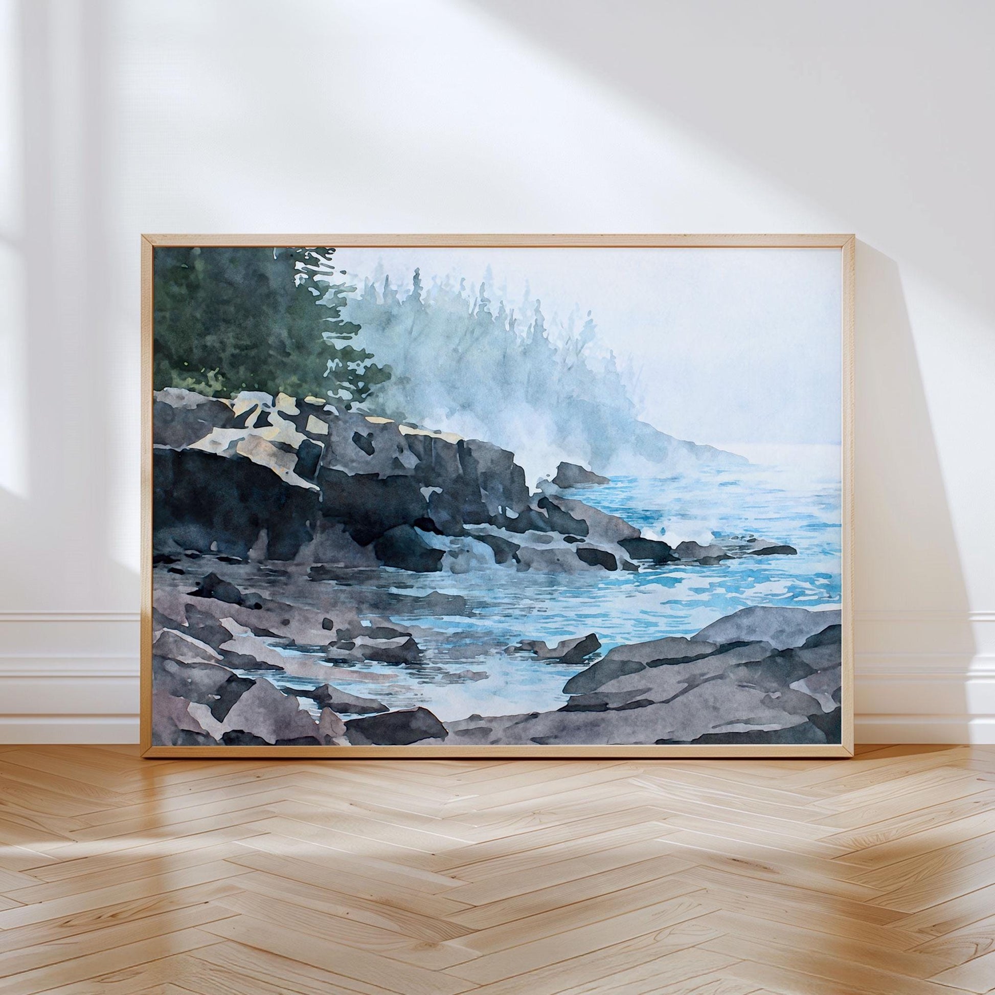 Large horizontal art print displayed in a light wood frame, leaning against a white wall on a hardwood floor. The artwork shows a watercolor-style misty Lake Superior shoreline with dark rocky cliffs in the foreground and calm blue water flowing between layered stones. Soft fog drifts across the scene, partially obscuring a distant evergreen forest, creating a serene, atmospheric lakeside mood in muted blues, grays, and greens.