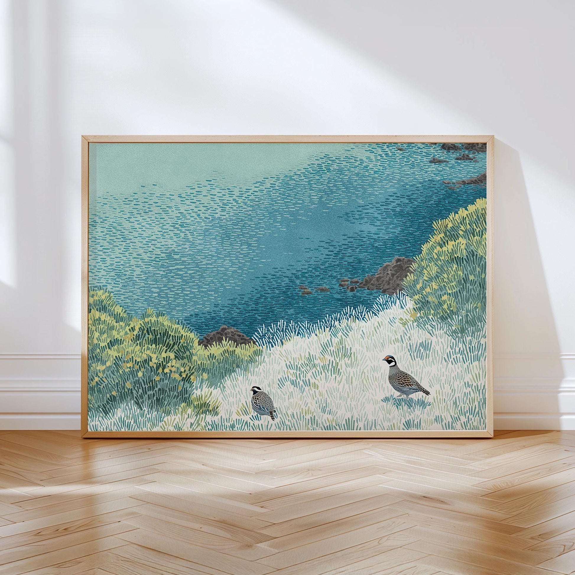 Framed art print featuring two California quail in a stylized coastal meadow, with textured grasses, vibrant green shrubs, and a rich blue ocean beyond. The piece is propped against a white wall on a light wood herringbone floor, with natural light enhancing its tranquil, nature-inspired palette.
