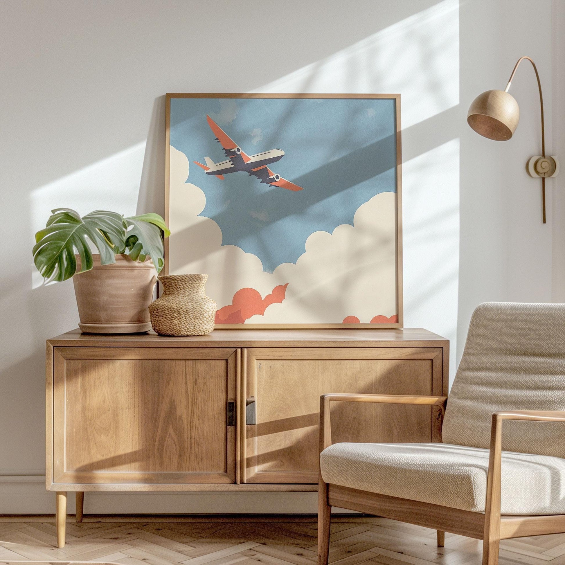 Framed airplane art print leaning against a white wall atop a wooden credenza, styled with a potted monstera plant, a woven vase, and bathed in warm sunlight; nearby is a modern wood and cream armchair beneath a brass wall sconce.