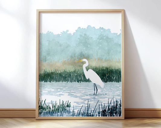 Minimalist watercolor-style art print of a white egret standing in shallow water surrounded by tall marsh grasses, with soft green trees and misty blue sky in the background. The artwork is framed in a light wood frame and leaning against a wall on a wooden floor.