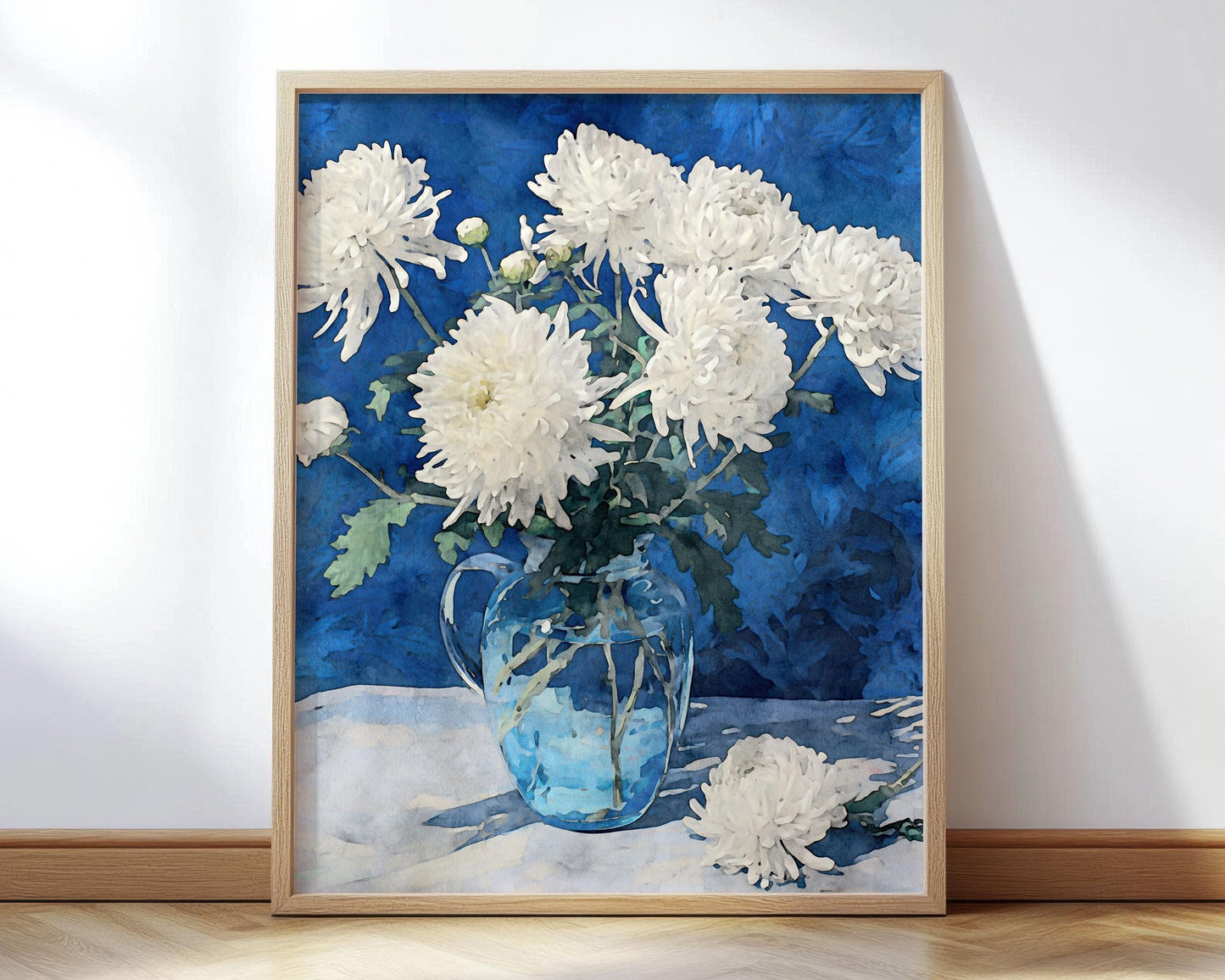 Framed art print of white chrysanthemums arranged in a clear glass pitcher, set against a deep blue background. One flower lies on the table beside the vase, creating a serene still life composition.