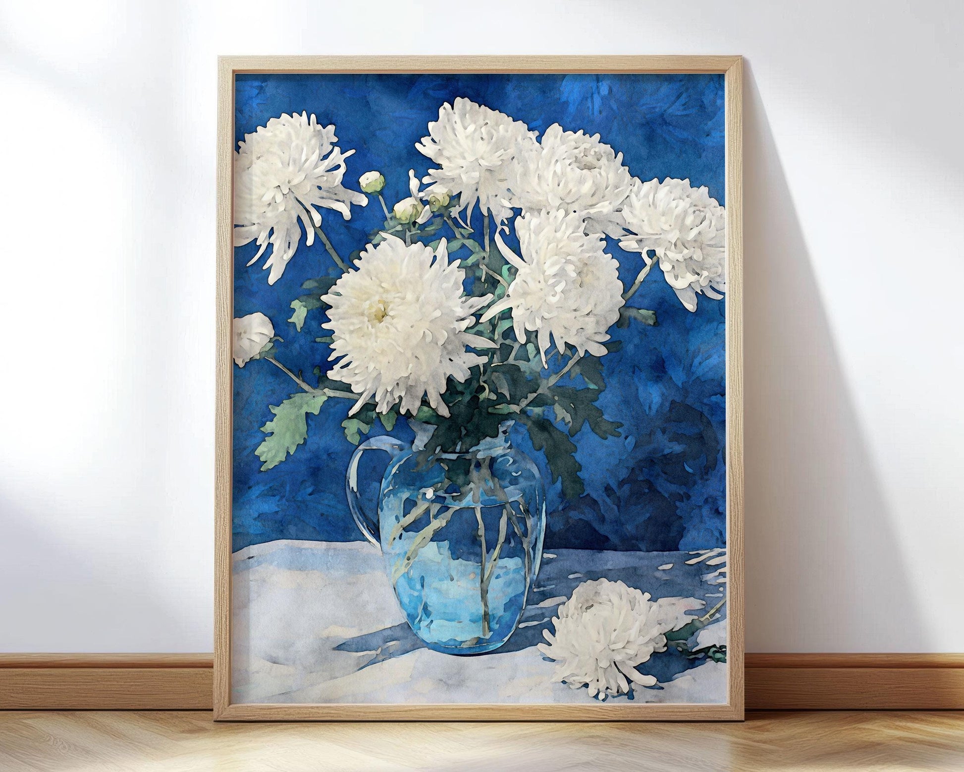 Framed art print of white chrysanthemums arranged in a clear glass pitcher, set against a deep blue background. One flower lies on the table beside the vase, creating a serene still life composition.