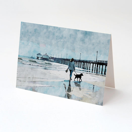 Greeting card featuring a watercolor-style illustration of a woman walking a black dog along the shoreline near a long pier, under a misty blue sky; card is displayed upright on a clean white surface.