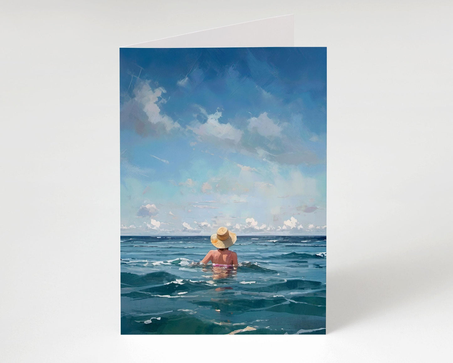 Front view of a standing greeting card showing a woman in a straw hat immersed in deep blue water under a wide sky with gentle clouds.