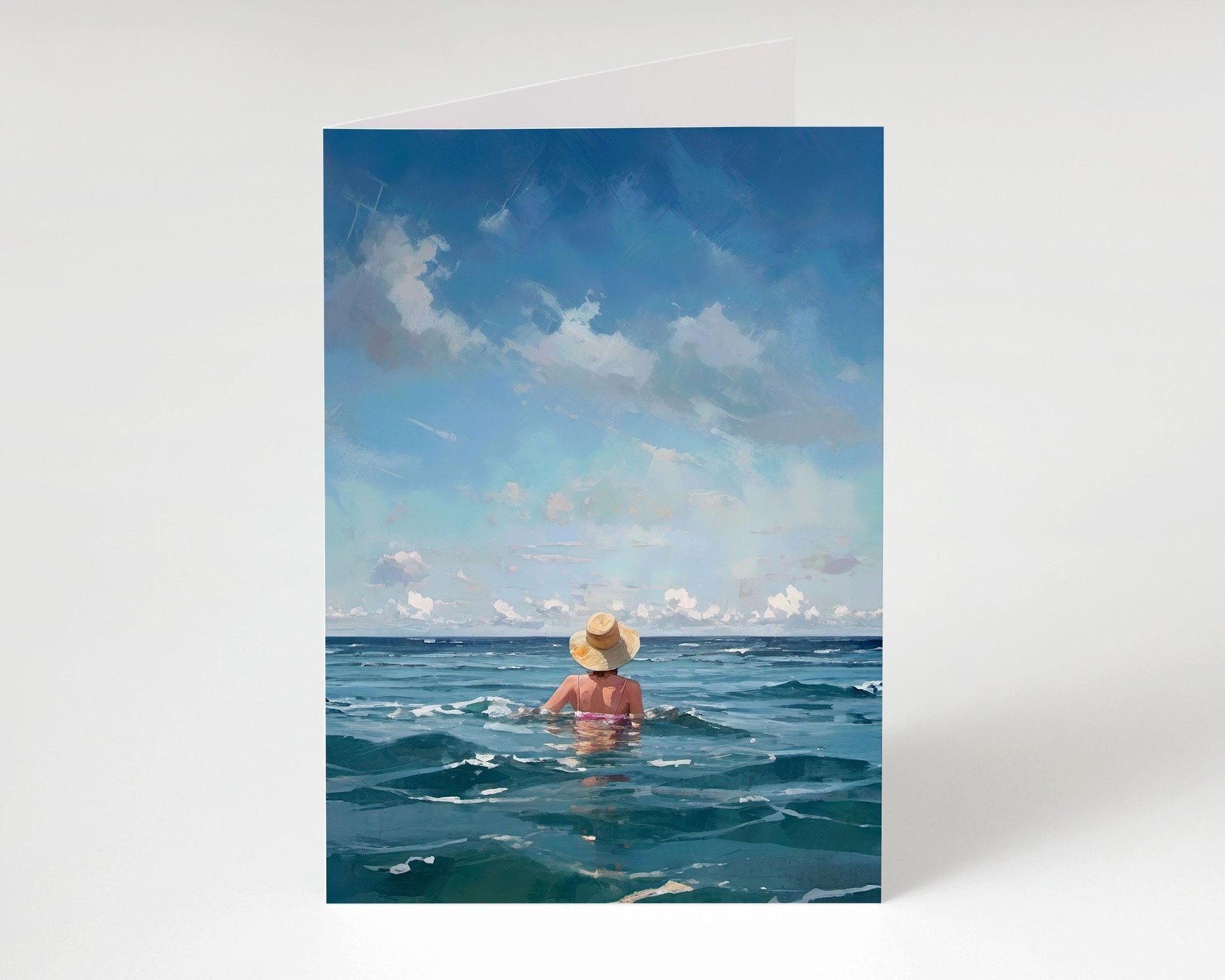 Front view of a standing greeting card showing a woman in a straw hat immersed in deep blue water under a wide sky with gentle clouds.