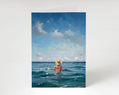 Front view of a standing greeting card showing a woman in a straw hat immersed in deep blue water under a wide sky with gentle clouds.