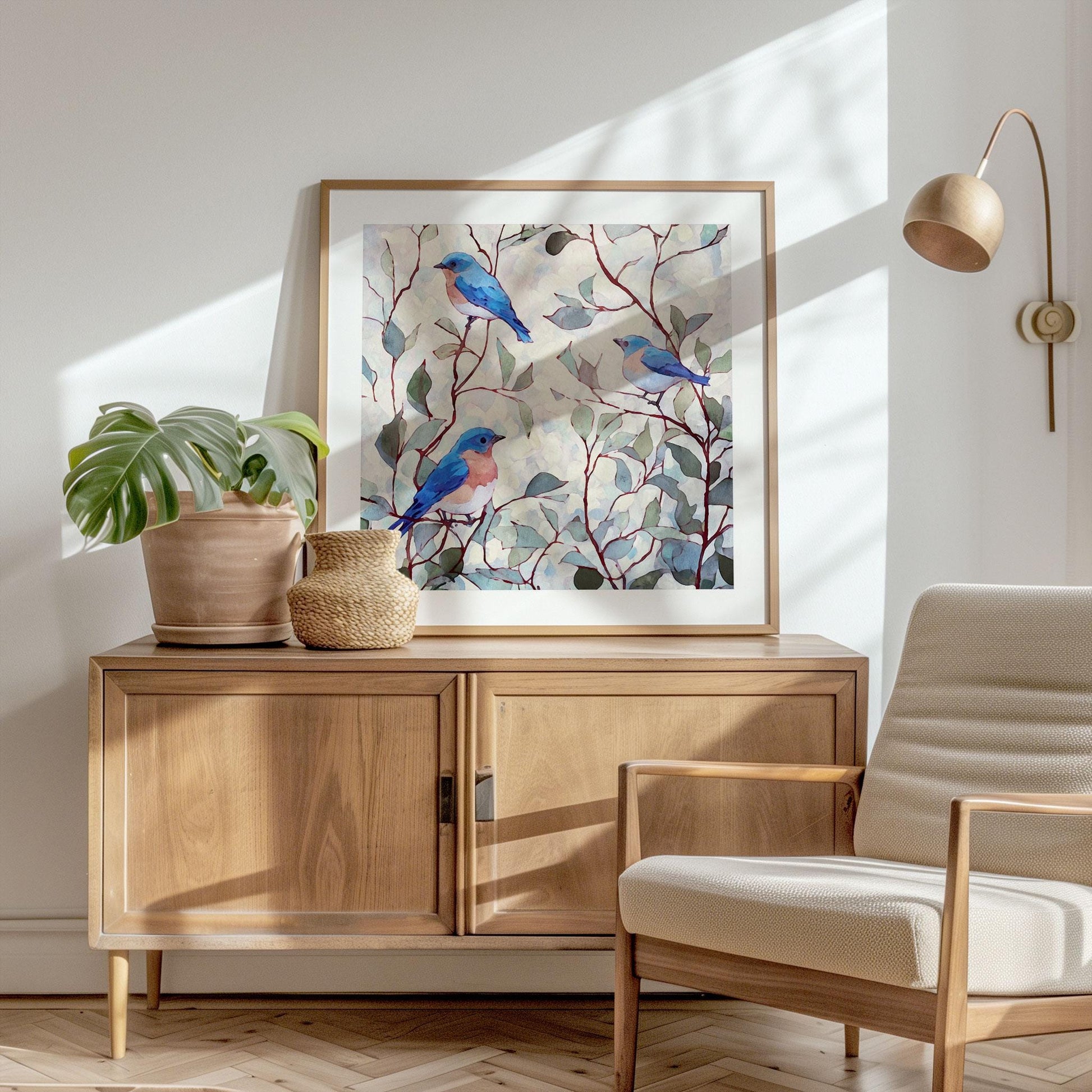 Framed watercolor art print of bluebirds perched on leafy branches, displayed on a wooden sideboard in a bright, minimalist living room. The artwork features soft blue and peach birds amid green and gray foliage on a pale background. A potted monstera and woven vase sit beside the frame, with a cream lounge chair and brass wall lamp completing the serene, Scandinavian-inspired space.