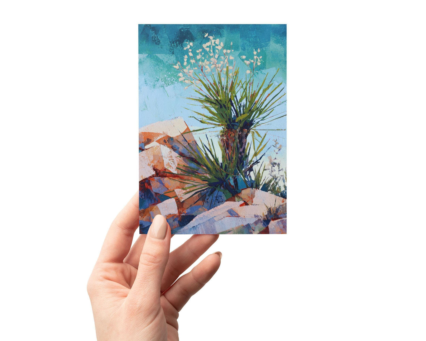 Hand holding a greeting card featuring a vibrant desert painting of a yucca plant with spiky green leaves and white blossoms, set among warm red and orange rocks beneath a bright blue sky.