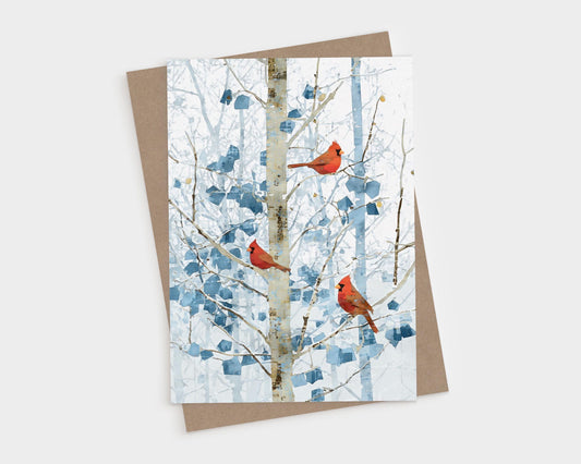 Greeting card featuring three bright red cardinals perched on slender branches of a winter tree. The background shows soft blue and white abstract foliage and a pale birch-like trunk, creating a serene snowy forest scene. Card is displayed on top of a kraft brown envelope against a light backdrop.