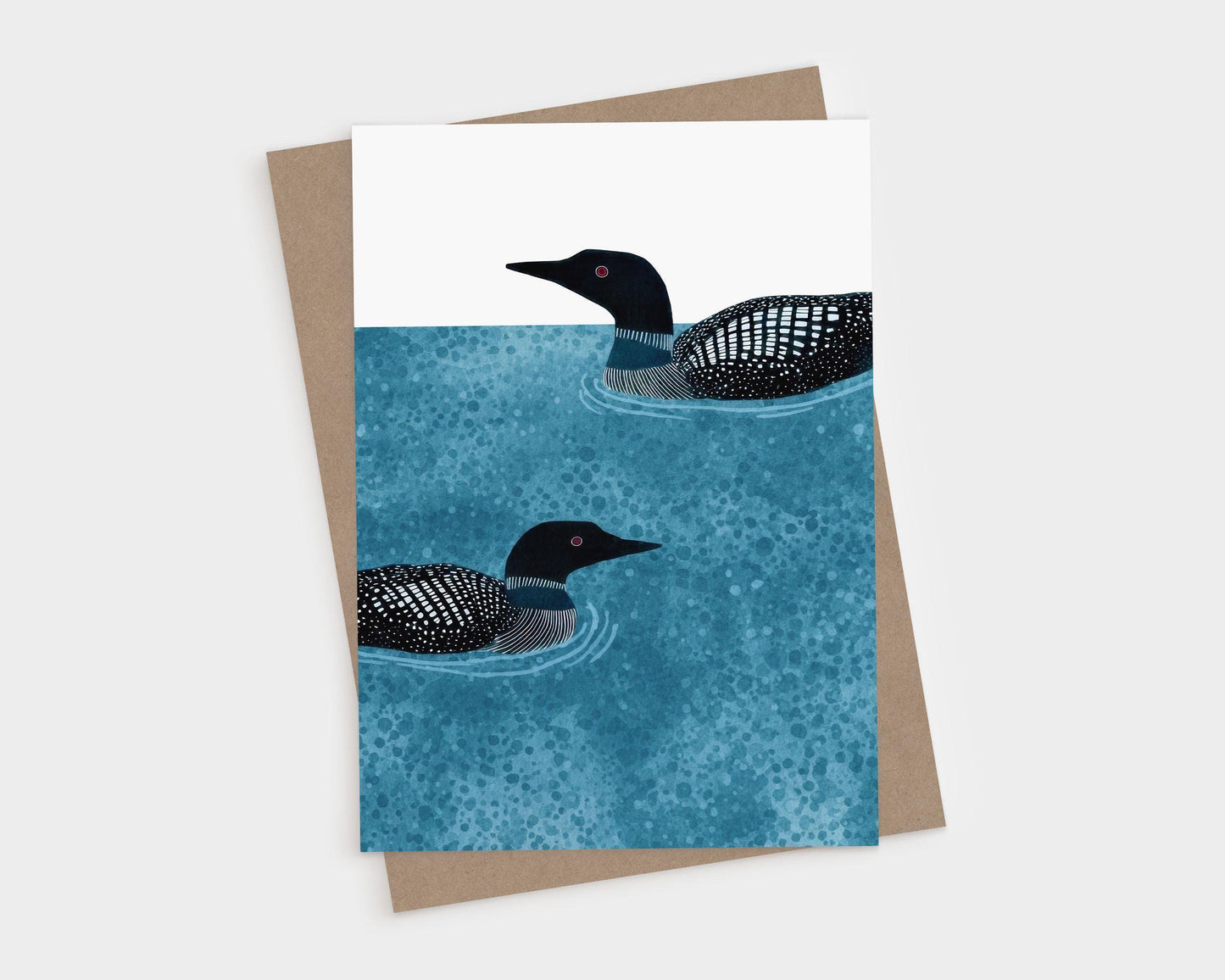 Vertical greeting card with two loons illustrated in black and white, swimming on textured deep blue water with ripples, set against a clean white background, paired with a kraft envelope.