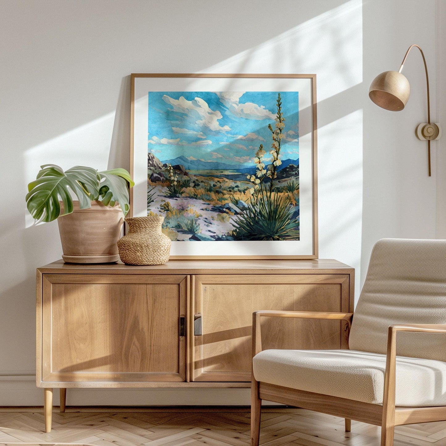 Framed desert landscape painting of yucca plants in bloom under a bright blue sky with scattered clouds, displayed on a wooden cabinet beside potted plants and a woven basket in a sunlit modern room with a lounge chair.