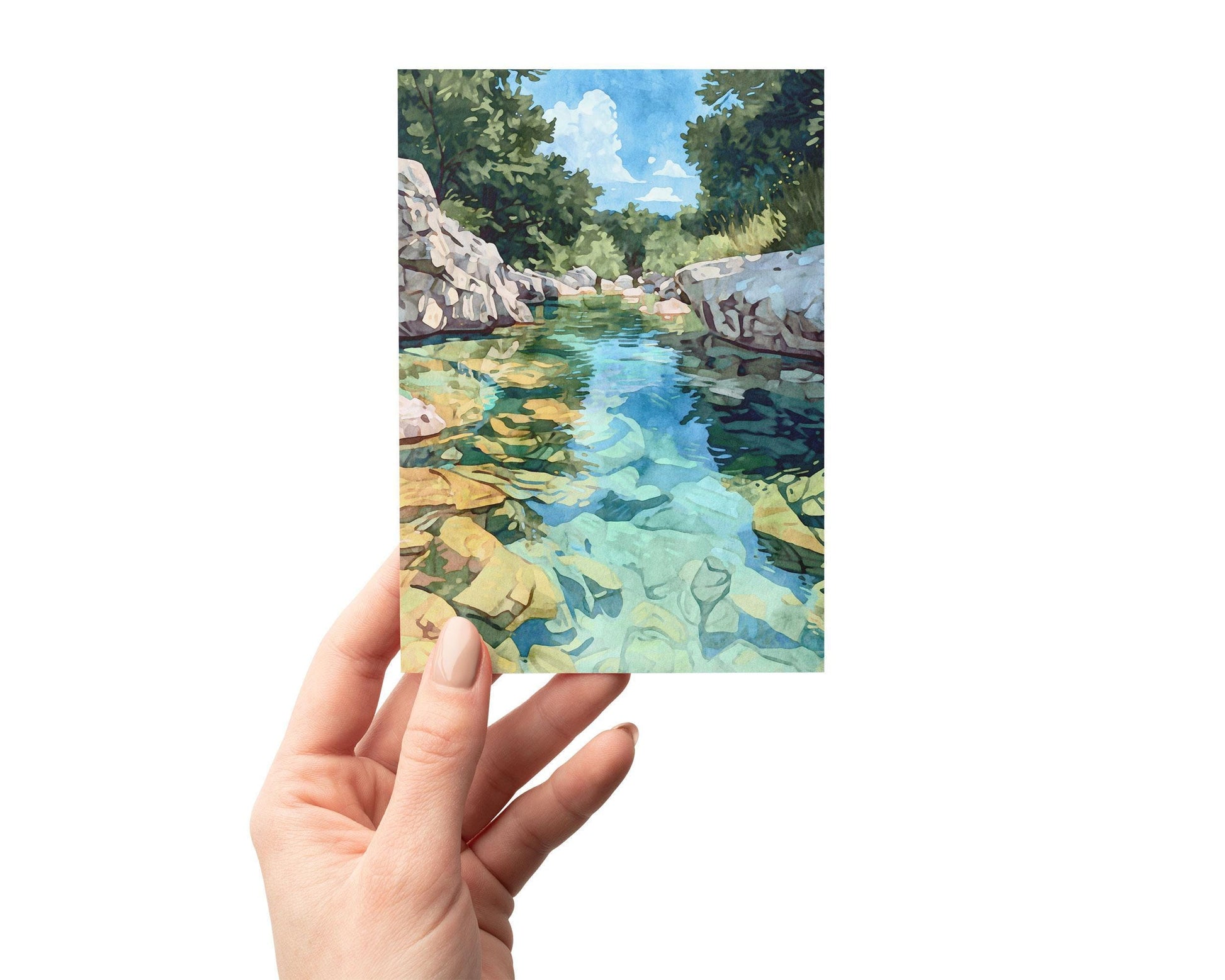 Hand holding a greeting card featuring a watercolor-style painting of a clear turquoise river with golden rocks visible beneath the water, framed by rocky banks and lush green trees under a bright blue sky.