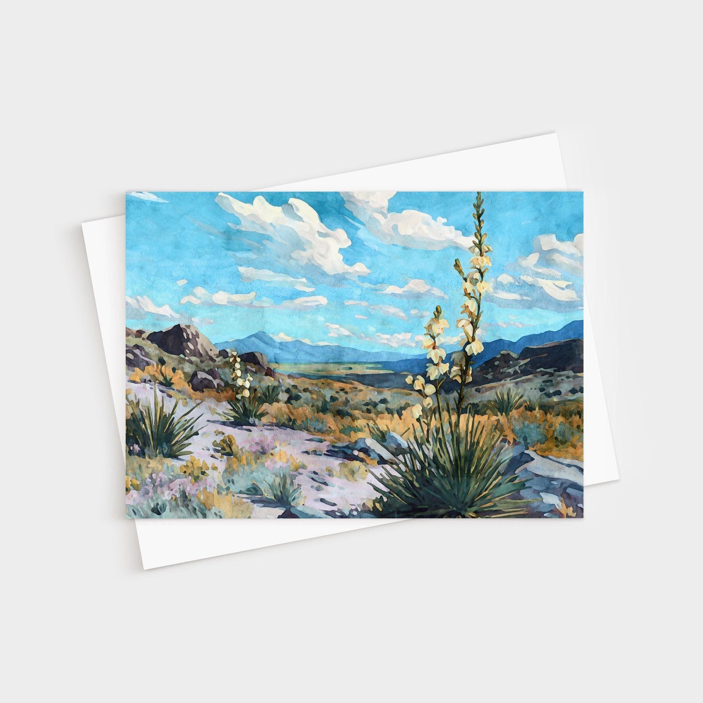 Greeting card featuring a watercolor-style desert landscape with blooming yucca plants, rocky hills, and distant mountains under a vivid blue sky with fluffy white clouds.