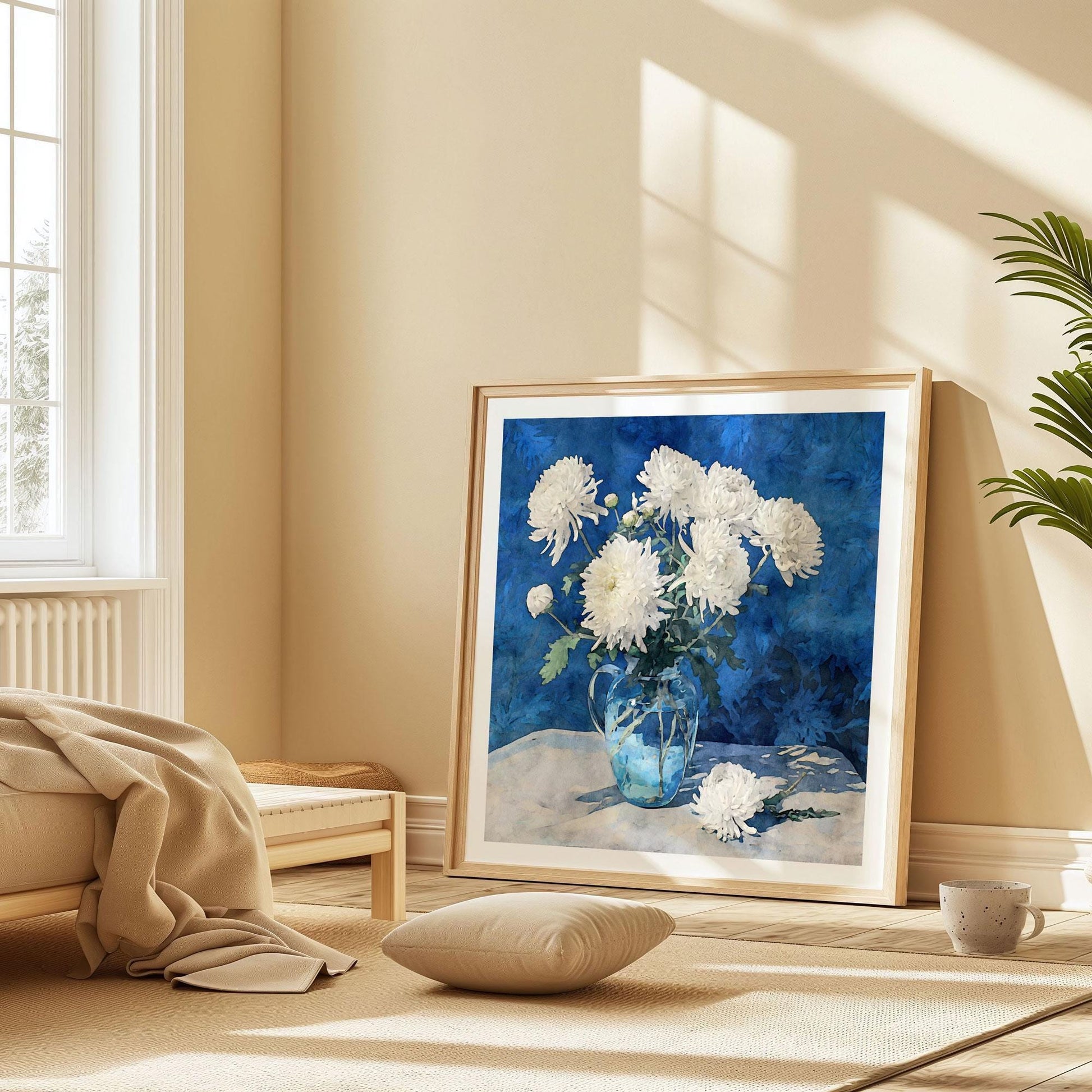 Large framed art print of white chrysanthemums in a glass pitcher with a rich blue background, leaning against a beige wall in a sunlit bedroom. The print rests on the floor beside a low bed with soft blankets, a cushion, and a ceramic mug, creating a cozy, relaxed atmosphere.