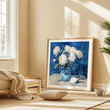 Large framed art print of white chrysanthemums in a glass pitcher with a rich blue background, leaning against a beige wall in a sunlit bedroom. The print rests on the floor beside a low bed with soft blankets, a cushion, and a ceramic mug, creating a cozy, relaxed atmosphere.