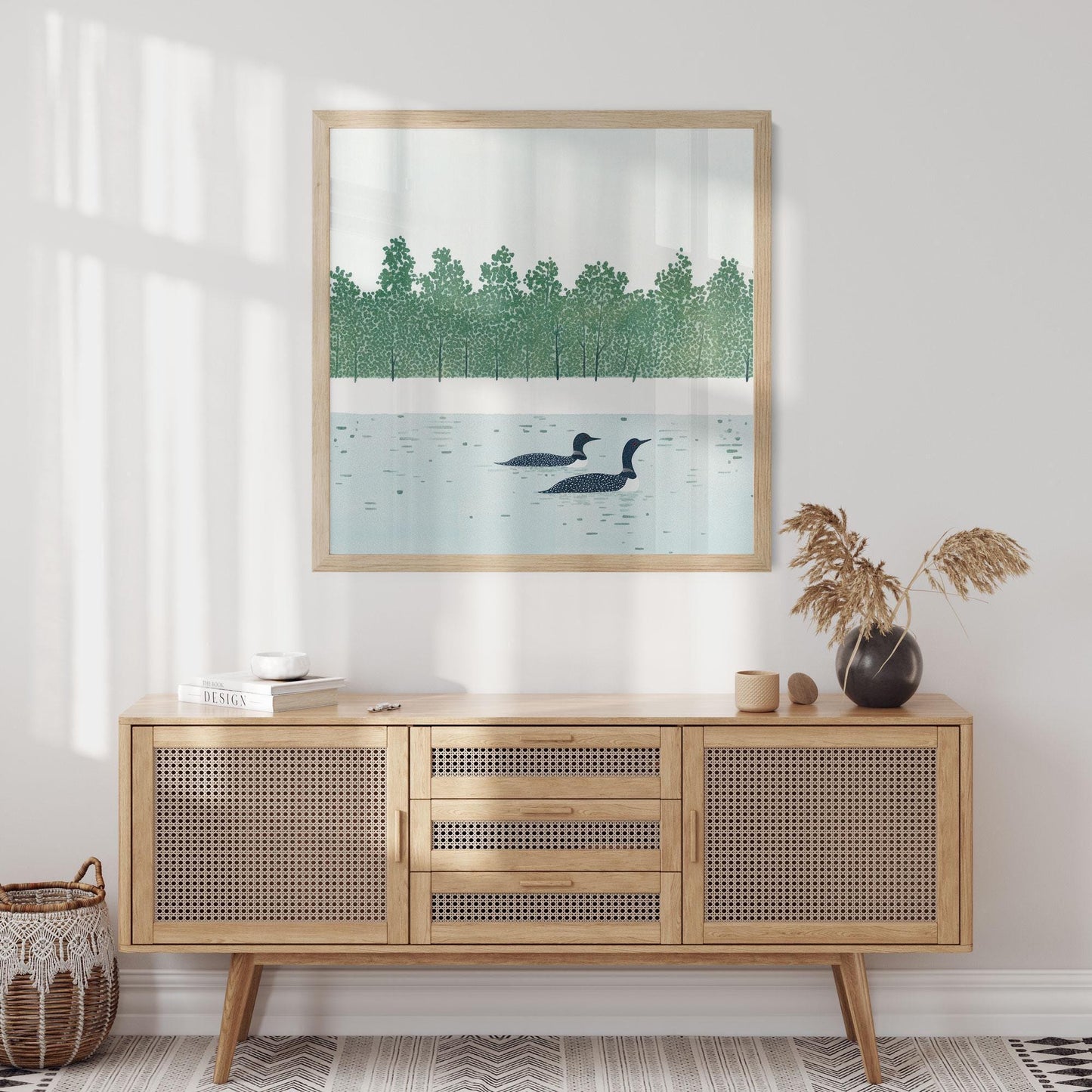 Framed loon art print displayed above a light wood cane-front sideboard in a minimalist room. The artwork depicts two loons floating on a still lake, with a stylized forest of green trees lining the far shore. Soft natural light filters in, highlighting neutral accents like a ceramic vase with dried grasses and a woven basket on the floor.