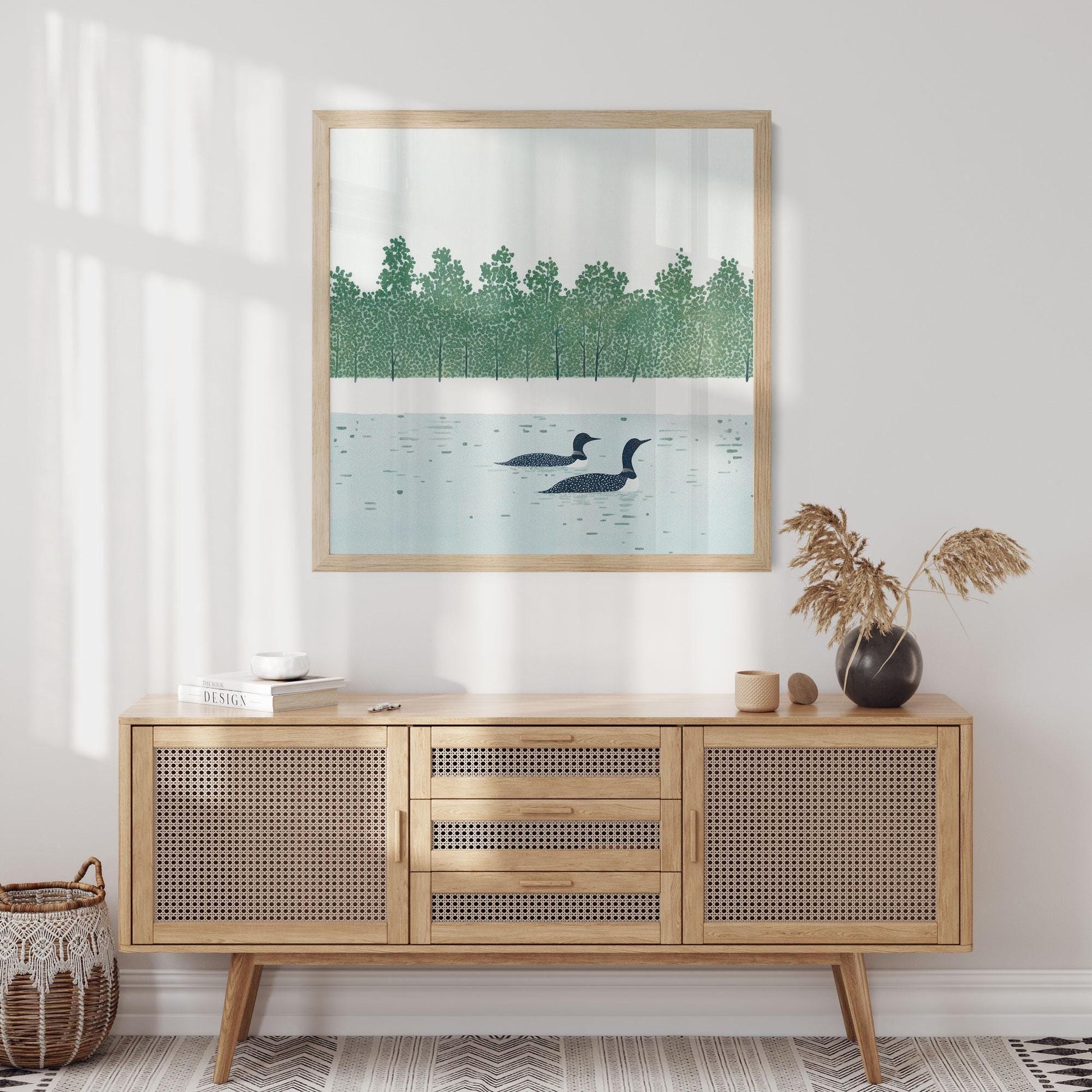 Framed loon art print displayed above a light wood cane-front sideboard in a minimalist room. The artwork depicts two loons floating on a still lake, with a stylized forest of green trees lining the far shore. Soft natural light filters in, highlighting neutral accents like a ceramic vase with dried grasses and a woven basket on the floor.
