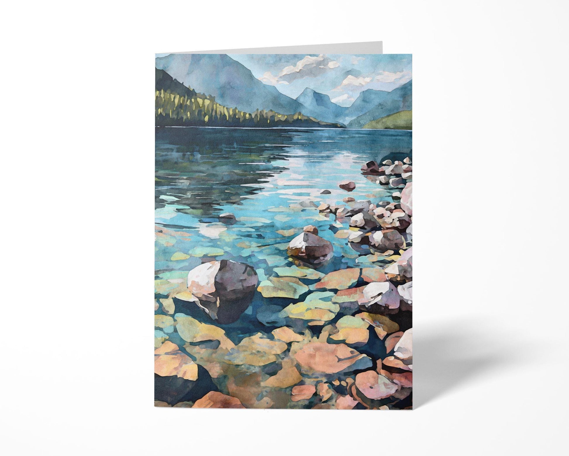 Standing greeting card featuring a watercolor-style illustration of a mountain lake with clear water revealing colorful rocks, a rocky shoreline, forested hills, and distant blue mountains beneath a partly cloudy sky.