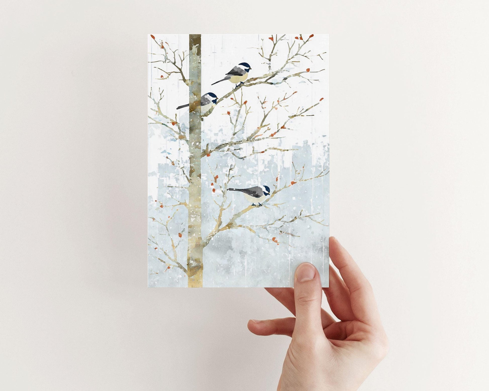 Hand holding a greeting card featuring a watercolor-style illustration of three chickadees perched on bare winter branches. The soft gray-blue snowy background and small orange berries create a calm, wintry look. The card is shown against a plain light background.