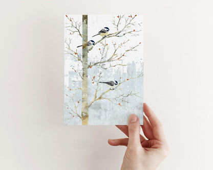 Hand holding a greeting card featuring a watercolor-style illustration of three chickadees perched on bare winter branches. The soft gray-blue snowy background and small orange berries create a calm, wintry look. The card is shown against a plain light background.
