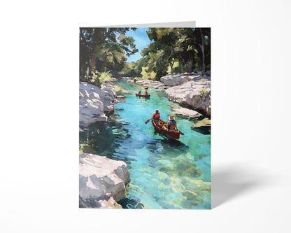 Folded greeting card featuring a watercolor-style painting of two red canoes gliding down a clear turquoise river, surrounded by rocky white banks and tall green trees under a bright blue sky. Displayed upright against a plain white background.