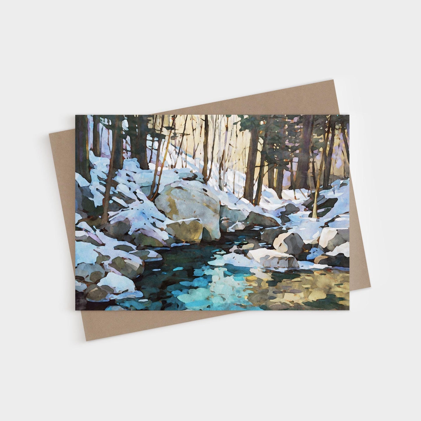 Greeting card featuring a watercolor-style painting of a snowy forest scene with large rocks and a creek reflecting icy blue and golden tones. The card is displayed with a kraft brown envelope underneath.