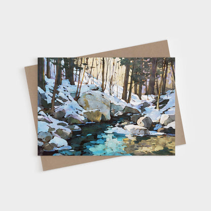 Greeting card featuring a watercolor-style painting of a snowy forest scene with large rocks and a creek reflecting icy blue and golden tones. The card is displayed with a kraft brown envelope underneath.