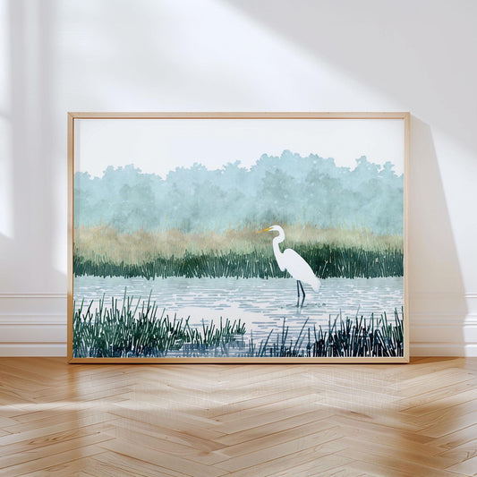 Framed watercolor-style print of a white heron standing in shallow water surrounded by tall grasses, with soft green trees and misty blue sky in the background, displayed leaning against a white wall on a wooden floor.