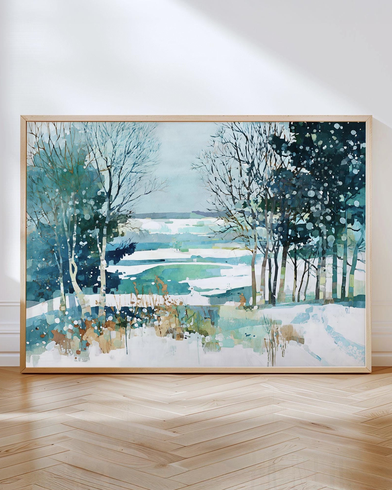 Framed watercolor landscape painting of a snowy winter scene with bare trees and evergreens overlooking a frozen lake in shades of teal, blue, and white, leaning against a white wall on a herringbone wood floor.