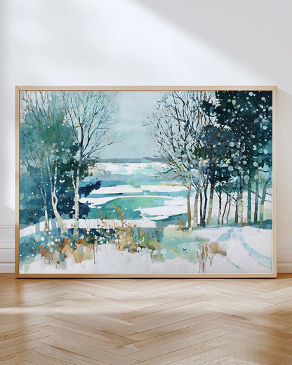 Framed watercolor landscape painting of a snowy winter scene with bare trees and evergreens overlooking a frozen lake in shades of teal, blue, and white, leaning against a white wall on a herringbone wood floor.