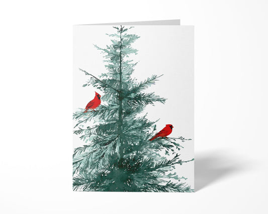 Folded greeting card standing upright on a white background, featuring a watercolor illustration of a pine tree with two bright red cardinals perched on its branches.