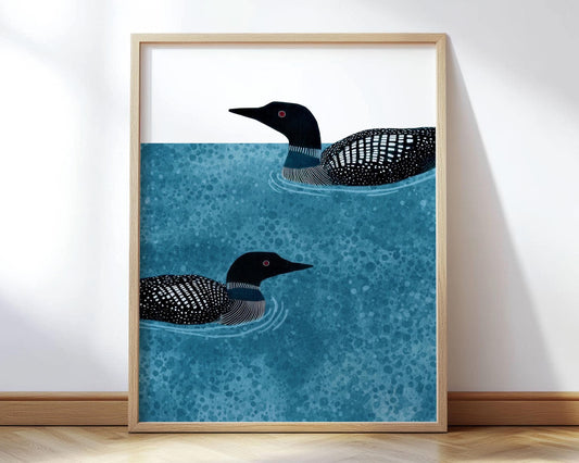 Framed vertical illustration of two loons swimming in mottled blue water, their black-and-white patterned feathers and red eyes contrasting with the crisp white background. The artwork rests against a white wall on a light wood floor, with natural light highlighting its clean, modern style.