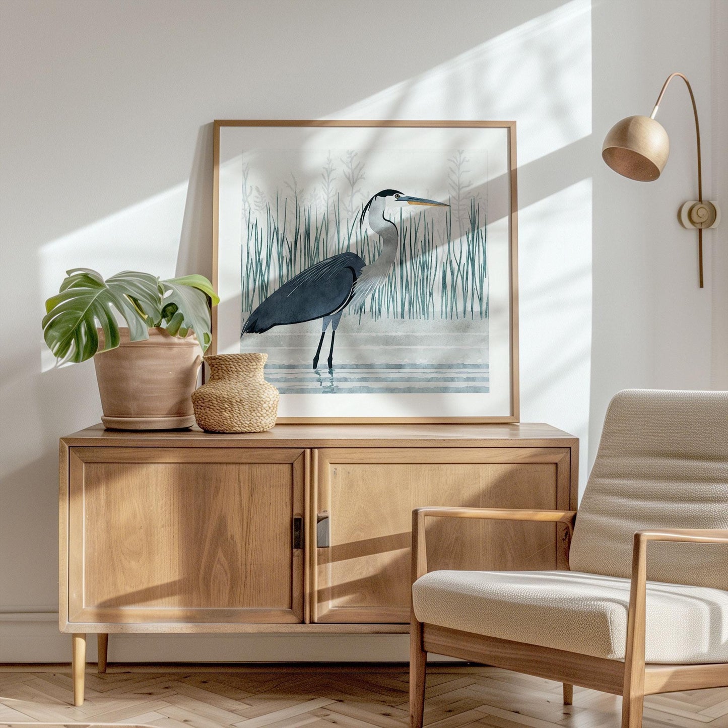 Framed art print of a great blue heron standing in water, resting on a light wood sideboard in a sunlit room. The heron is illustrated in a soft, watercolor style with tall reeds in the background. A potted monstera plant and woven vase sit beside the artwork, and a modern cream-colored armchair and wall-mounted lamp add to the room’s cozy, Scandinavian-inspired decor. Sunlight casts soft shadows across the white wall and parquet floor.