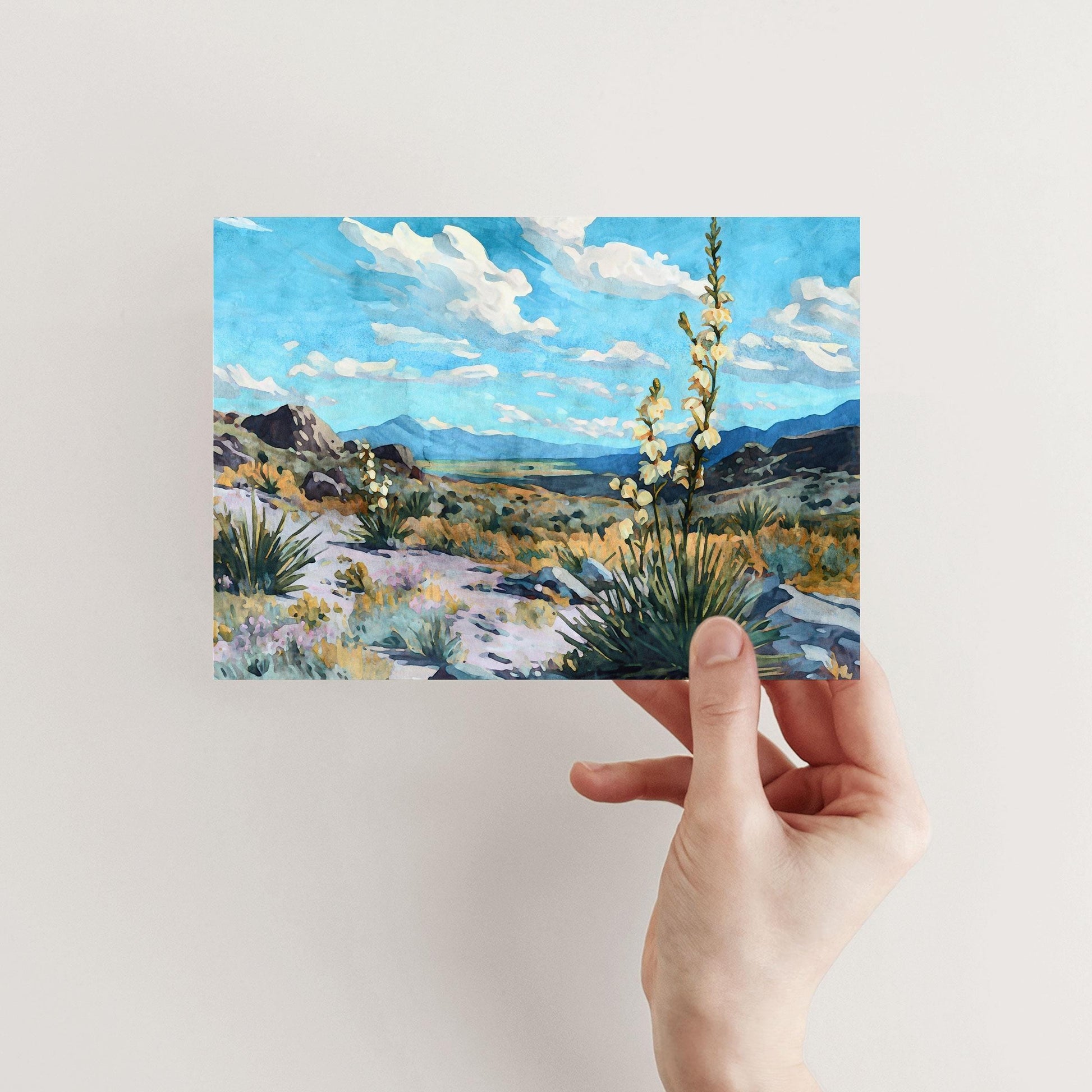 Hand holding a greeting card featuring a watercolor-style desert landscape with blooming yucca plants, rocky terrain, and distant mountains under a vivid blue sky with fluffy clouds.