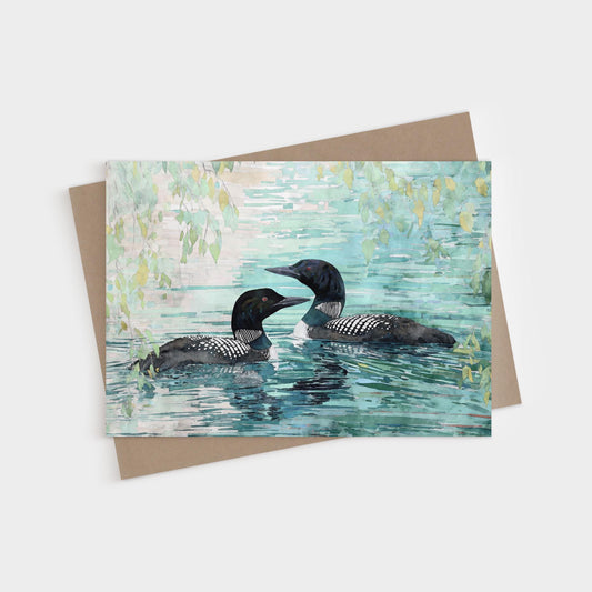 Watercolor greeting card featuring two common loons swimming side by side in a calm lake with reflections of green leaves and soft blue water, paired with a kraft envelope.