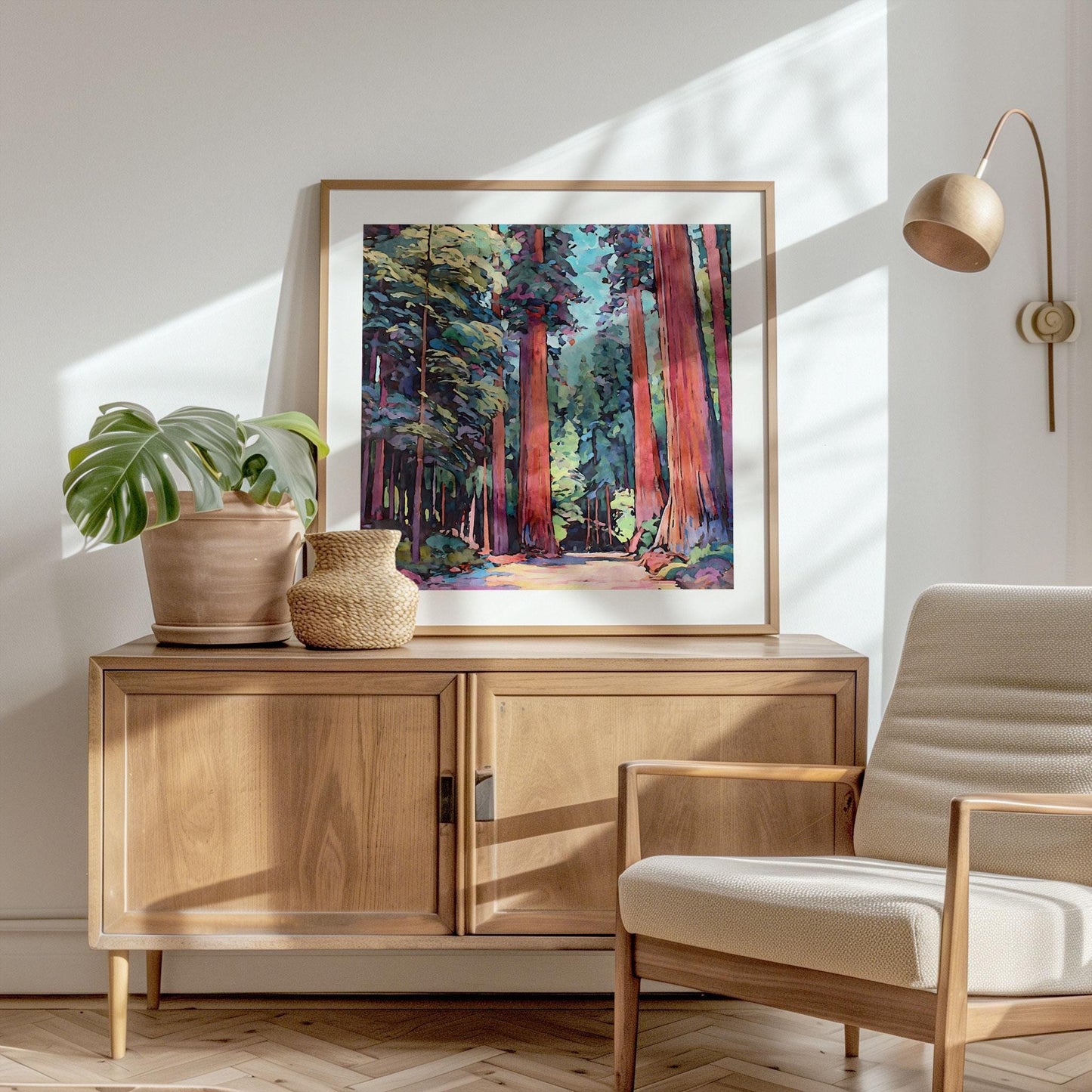 Framed art print of colorful impressionist-style redwood trees rests on a wooden sideboard in a bright, minimalist room. A potted monstera plant and woven vase sit beside the artwork, with a neutral armchair and wall-mounted lamp completing the modern, natural décor.