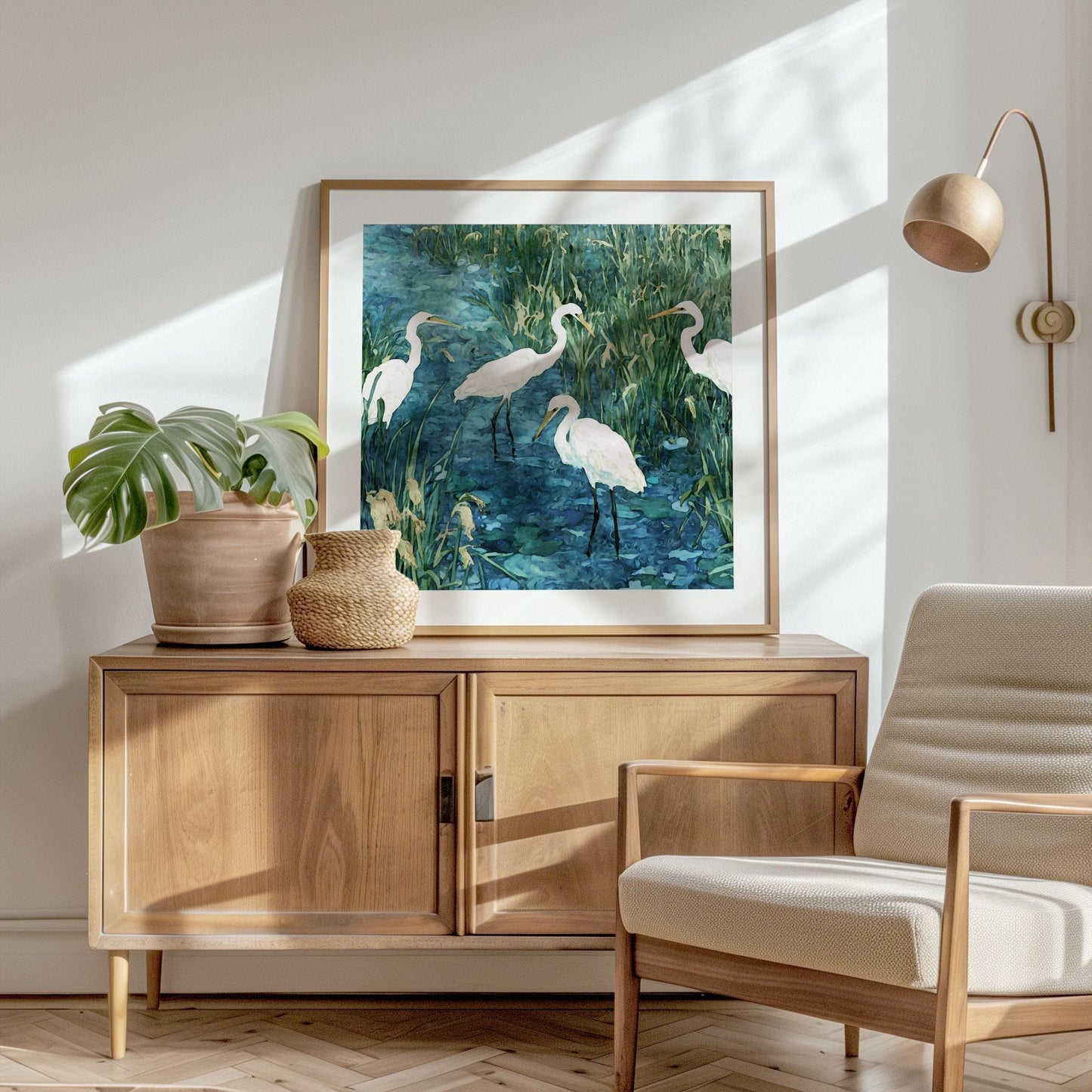 Square-framed painting of five white egrets standing in a marsh with tall grasses and blue water, resting on a wooden sideboard. A potted monstera plant and woven basket sit beside it, while a modern wood-and-fabric armchair and wall lamp complete the bright, sunlit minimalist room.