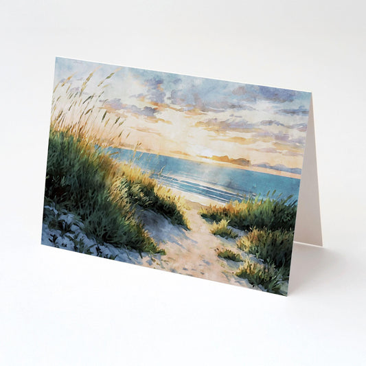 Folded greeting card featuring a watercolor painting of a sandy beach path through tall grasses leading to the ocean at sunset, with golden light reflecting on the waves and sky, displayed against a clean white background.