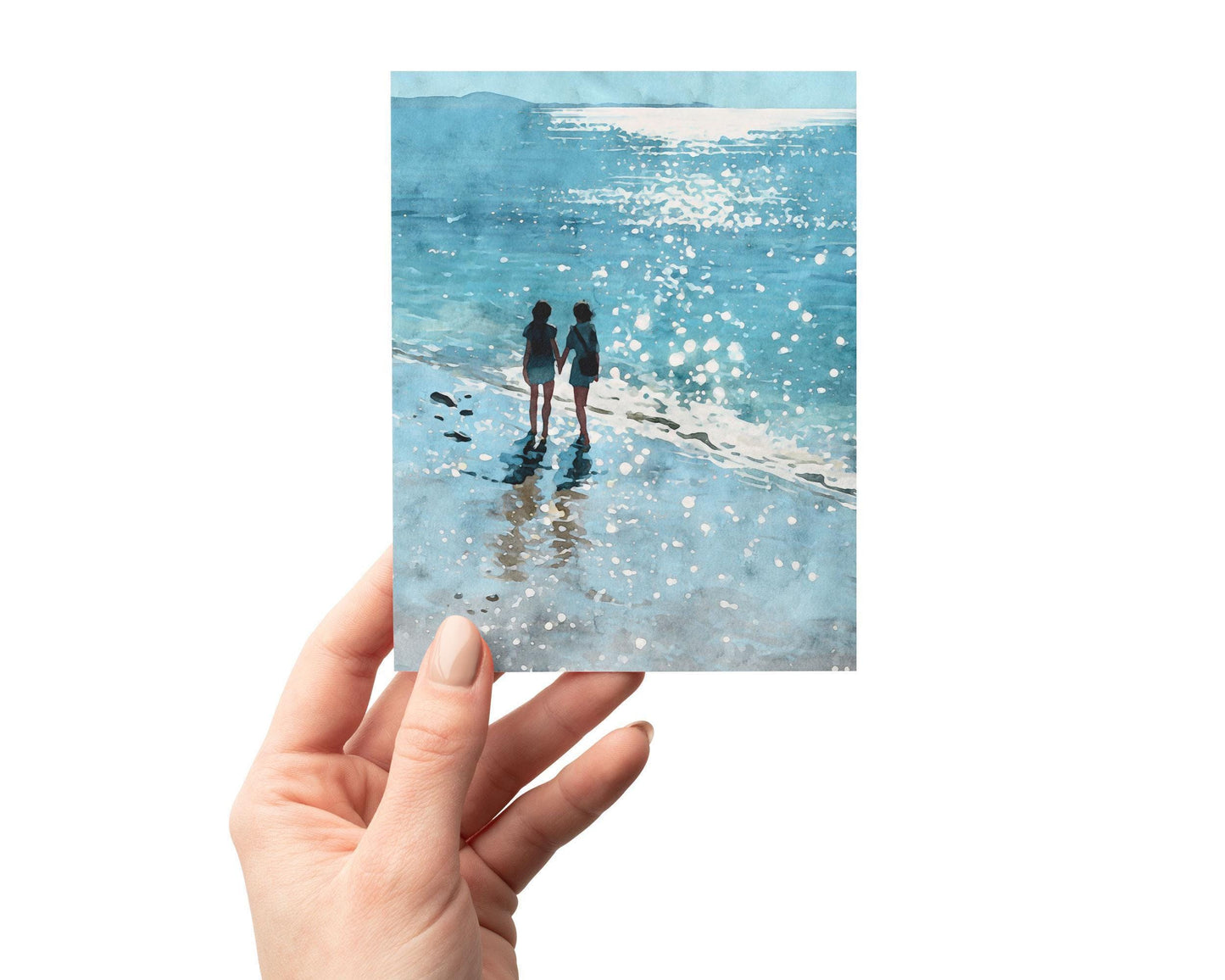 A hand holds a greeting card featuring a watercolor-style illustration of two people walking hand-in-hand along a shimmering beach, with sunlight sparkling on the water and their reflections mirrored in the wet sand.