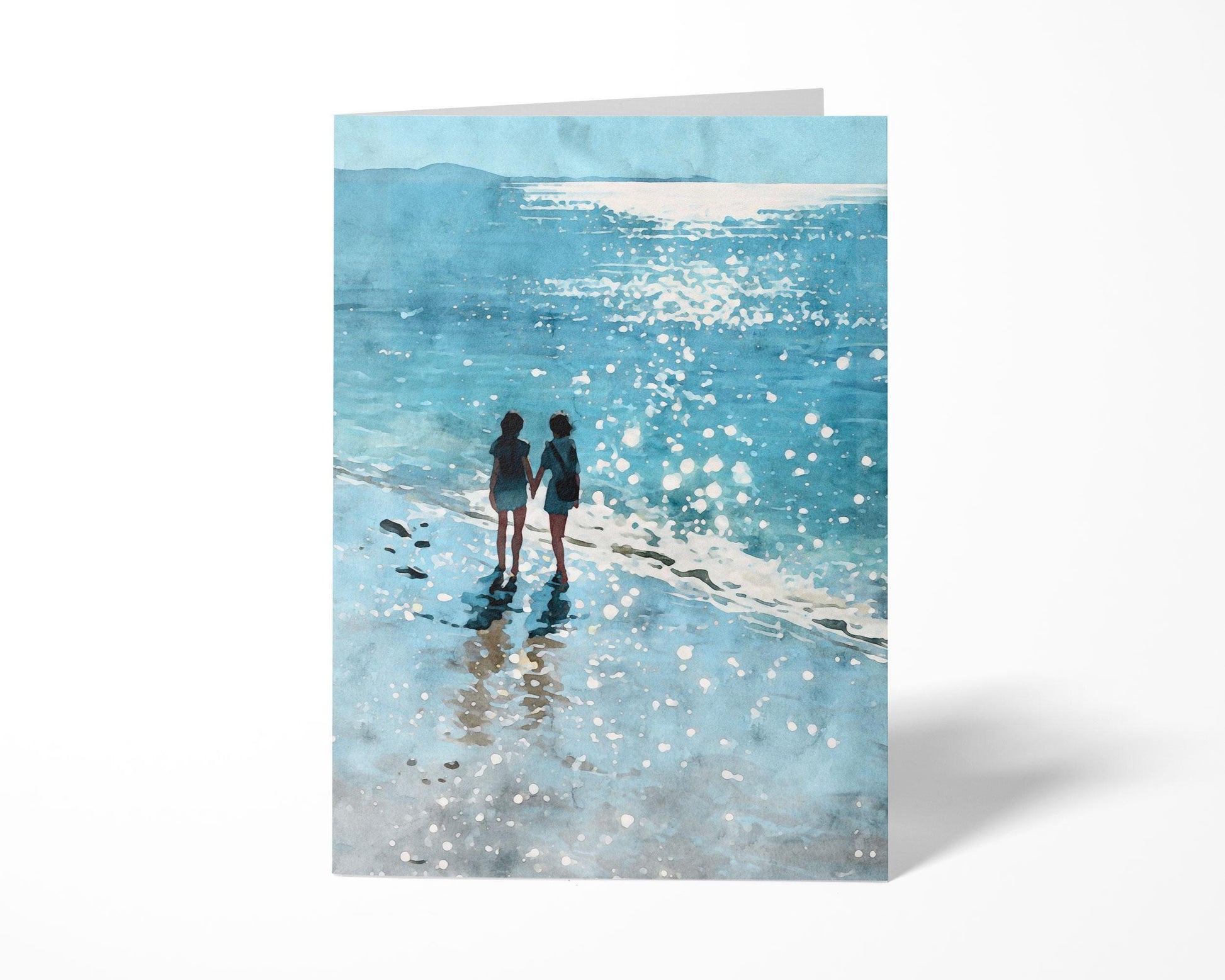 Greeting card standing upright against a white background, featuring a watercolor-style illustration of two people holding hands along a sunlit shoreline, with sparkling reflections on the ocean and wet sand.