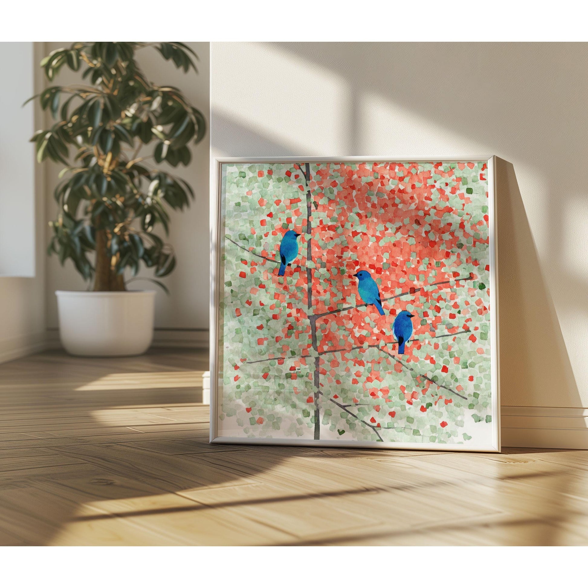 Framed art print of three vivid bluebirds perched on delicate branches of a maple tree with soft red and green foliage, painted in a mosaic watercolor style. The artwork rests on a wood floor near a sunlit wall, with a large potted plant in the background and warm natural light filling the minimalist room.