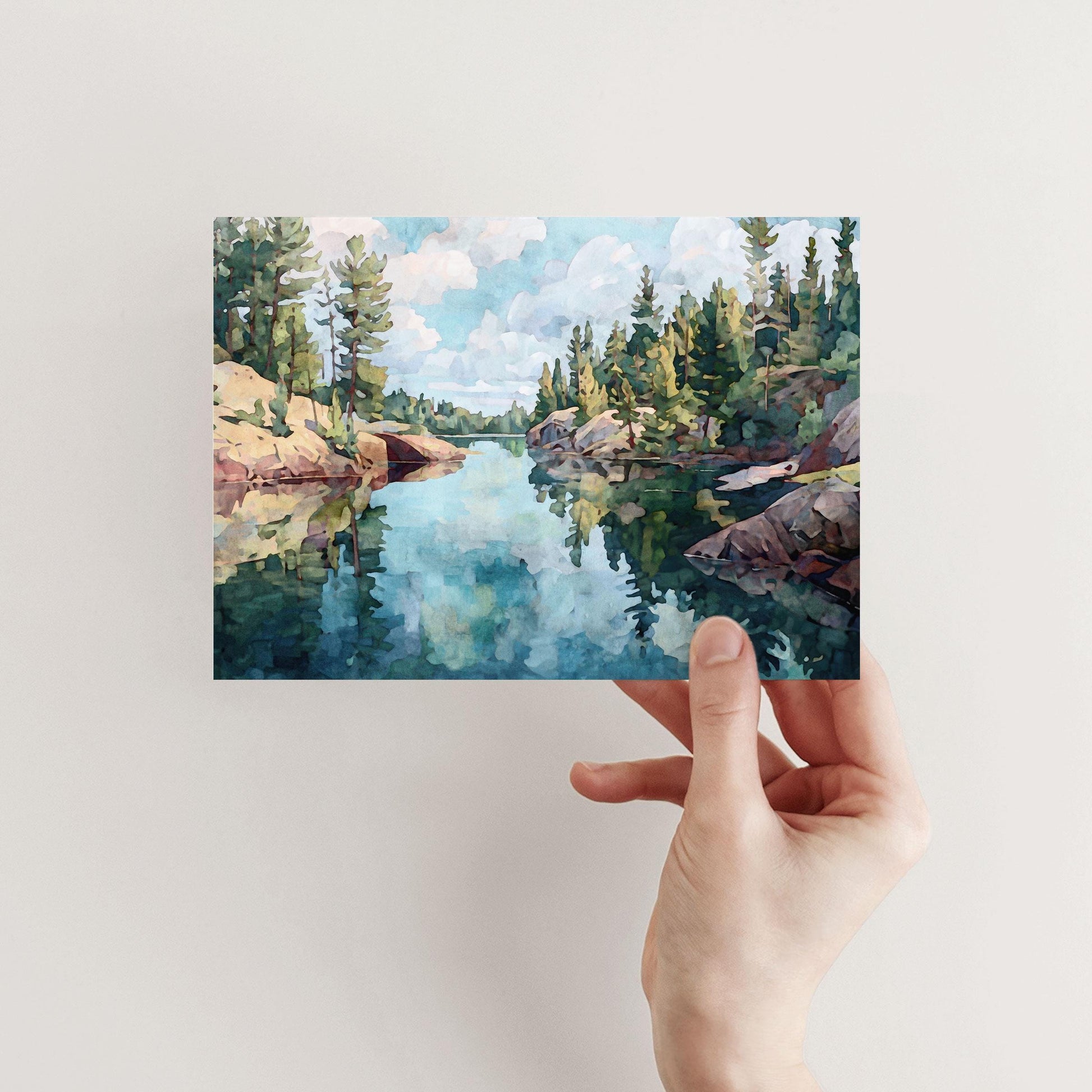 Hand holding a horizontal greeting card featuring a watercolor-style painting of a calm blue lake with rocky shores and evergreen trees, reflecting the sky and clouds in the still water.