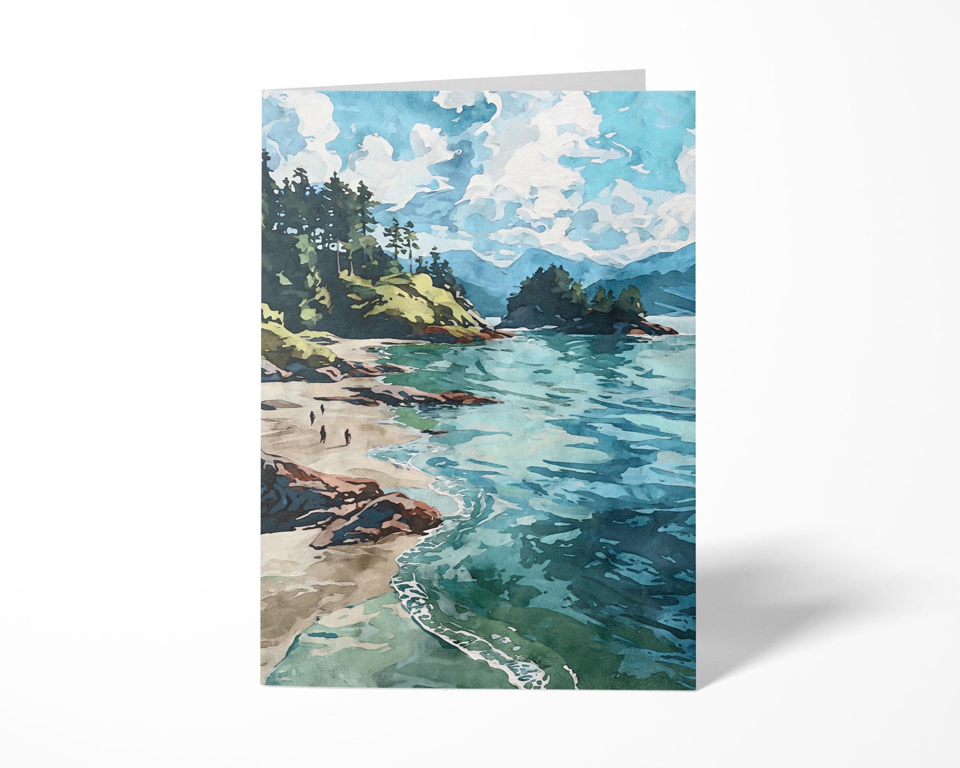 Greeting card standing upright on a white surface, featuring a watercolor-style coastal landscape with turquoise water, sandy beach, rocky cliffs, and pine-covered hills beneath a sky filled with fluffy white clouds.