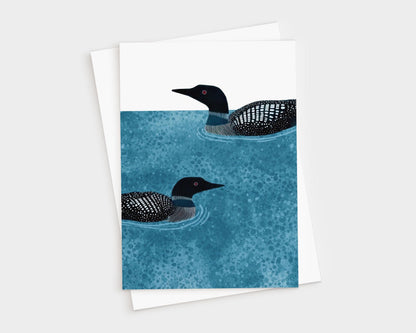 Vertical greeting card with two loons illustrated in black and white, swimming on textured deep blue water with ripples, set against a clean white background, paired with a white envelope.
