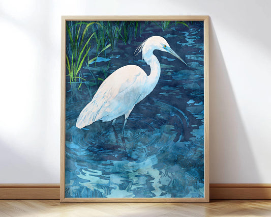 Framed art print of a white egret standing in rippling blue water with reflections, surrounded by tall green reeds, rendered in a watercolor style with soft, layered shades of blue and teal.