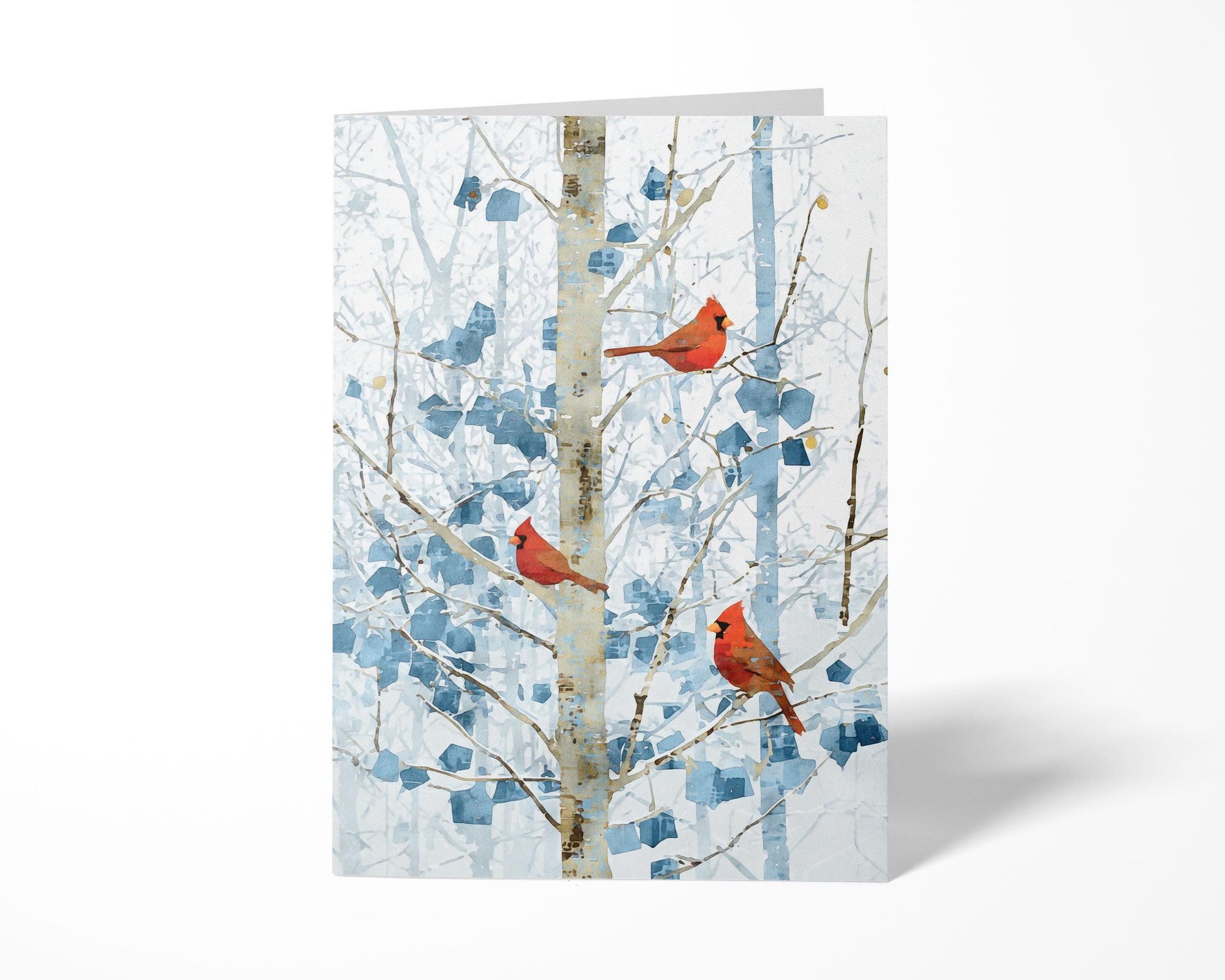 Folded greeting card featuring three bright red cardinals perched on pale winter branches with abstract blue leaves. The card stands upright against a clean white background, casting a soft shadow to the right.