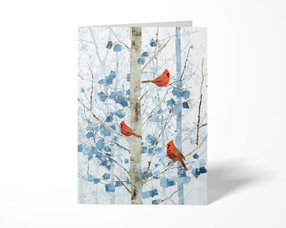 Folded greeting card featuring three bright red cardinals perched on pale winter branches with abstract blue leaves. The card stands upright against a clean white background, casting a soft shadow to the right.