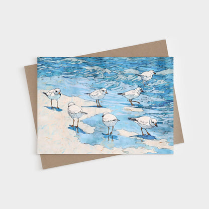 Watercolor-style illustration of small white shorebirds with orange legs standing and wading at the edge of blue ocean waves on a sandy beach, printed on a folded greeting card with a kraft brown envelope behind it.