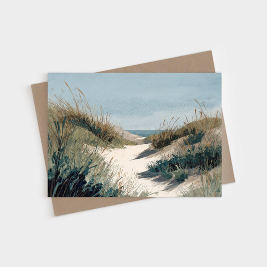 Greeting card featuring a serene watercolor illustration of a sandy path winding through grassy dunes toward the ocean, under a soft blue sky. A kraft paper envelope is placed behind the card.