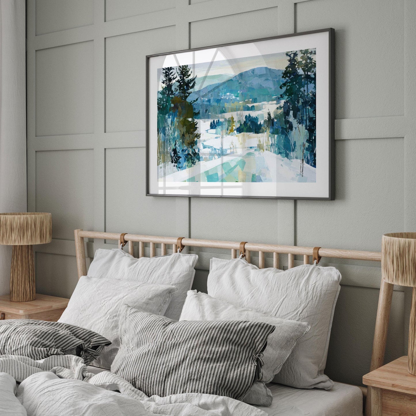 Framed winter landscape painting featuring snow, evergreen trees, and blue-green mountains, displayed above a bed with white linens and striped pillows in a modern, neutral-toned bedroom with wood accents and soft natural light.